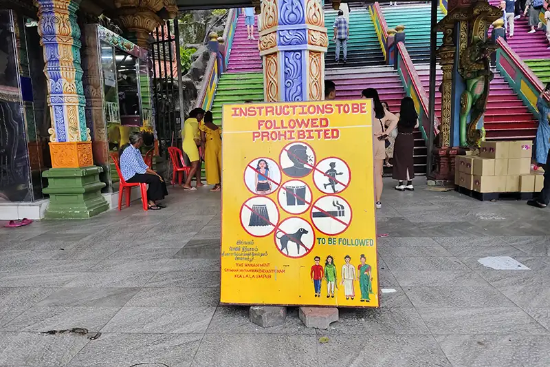 Signboard displaying the dress code and prohibited actions for visitors at Batu Caves, Kuala Lumpur, near the colorful staircase.