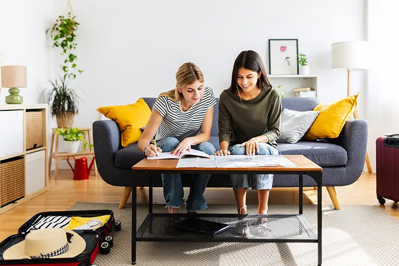 Two young women planning their trip to Malaysia, sitting in a cozy living room with travel guides and open luggage, emphasizing budget travel preparation.
