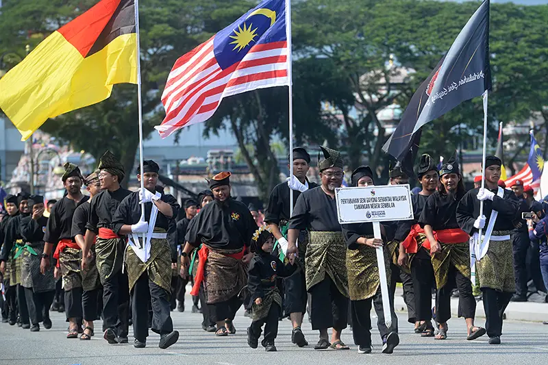 Cultural parade during Malaysia National Day celebrations with participants dressed in traditional attire carrying flags.