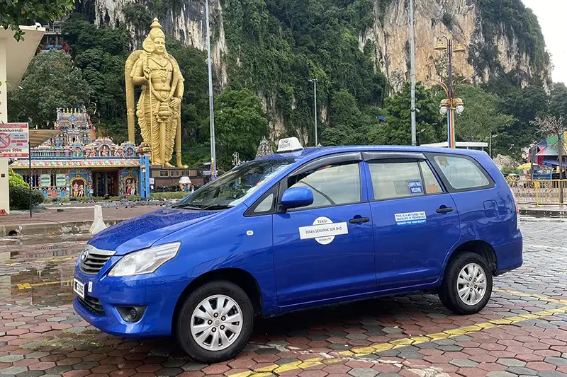 Blue taxi parked at Batu Caves with the golden Lord Murugan statue in the background.