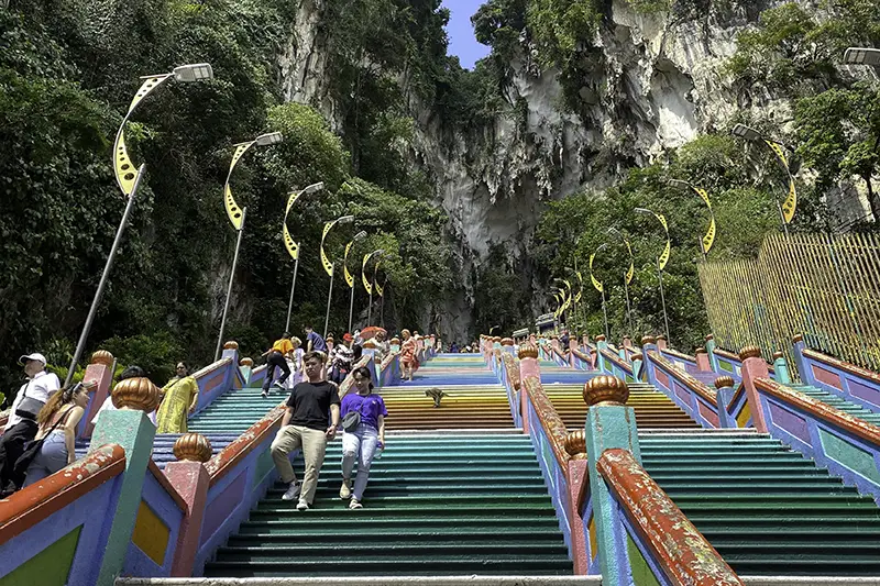 Tourists climbing the Batu Caves staircase in the late afternoon, with clear skies and soft lighting enhancing the colorful steps and limestone cliffs.
