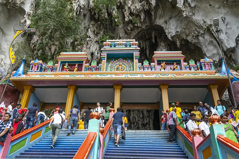 Tourists standing near the colorful archway at the top of the Batu Caves staircase, next to the main entrance of the upper cave temple.