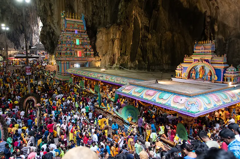 Massive crowd inside Batu Caves upper cave during Thaipusam festival, with colorful temple structures and Hindu devotees in procession.